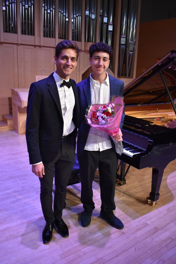 Two students standing together on stage after a recital, one holding a bouquet of flowers